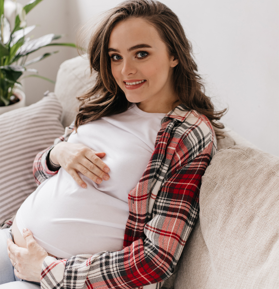 Womens & Maternal Health - Blue-eyed brunette pregnant woman sincerely smiles and looks into camera. Charming lady in plaid shirt touches belly and poses on soft sofa.